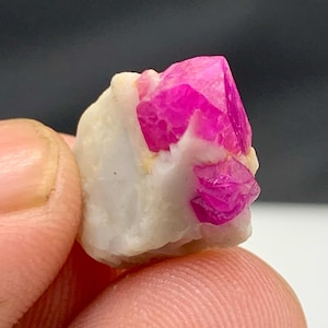 May include: A close-up of a small, raw mineral specimen. The mineral features a vibrant, hot pink crystal formation atop a white, rough matrix. The crystal has sharp, defined edges and a glossy appearance.