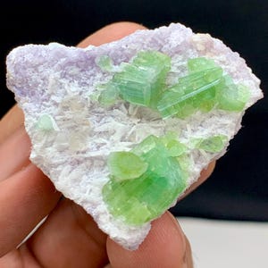 Może przedstawiać: Naturalny okaz minerału z żywymi zielonymi kryształami na matrycy z jasnofioletowego i białego kamienia. Zielone kryształy mają strukturę pryzmatyczną, kontrastującą z bardziej miękką, teksturowaną podstawą. Unikalny przedmiot dekoracyjny.