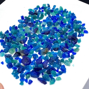 Puede incluir: Una colección de pequeños cristales de forma irregular en tonos azules y verdes. Los cristales están dispersos sobre una superficie blanca, creando una exhibición visualmente atractiva de color y textura. Las diferentes tonalidades sugieren diferentes composiciones minerales.