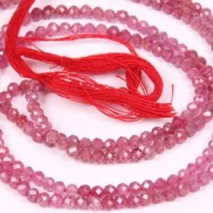 May include: Strands of faceted pink gemstones, likely tourmaline or similar, are displayed against a white background. A red string is attached to the gemstones. The beads are small and uniform in size, creating a delicate appearance.