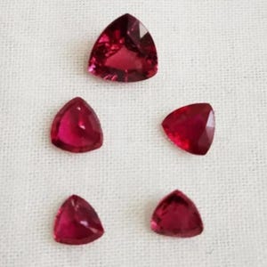 May include: Five triangular, faceted, deep red gemstones arranged on a white, textured surface. The gems are cut with flat backs and pointed tips, showcasing their vibrant color and clarity. The arrangement suggests a star or floral pattern.