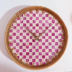 May include: Round wall clock with a woven rattan frame. The clock face features a checkered pattern of pink and white squares. The clock hands are made of wood. A decorative piece for any room.