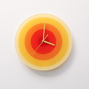 May include: A round wall clock with a retro design. The clock face features concentric circles in shades of yellow, orange, and red. The clock hands are gold-colored. The clock is a decorative item.