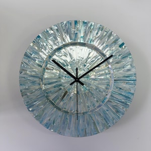 May include: A circular wall clock featuring a mosaic face composed of iridescent, light blue and white shell-like shards arranged in a radiating sunburst pattern. The clock has three thin, black hands positioned at the center of the dial.
