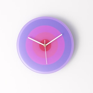 Peut inclure: Une horloge murale ronde avec un motif concentrique dans les tons de violet, rose et rouge. Le cadran de l'horloge présente un motif dégradé, avec un centre rouge et des aiguilles blanches. L'horloge est un objet décoratif.