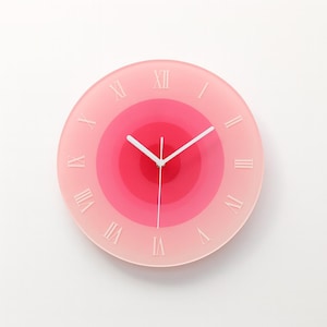 May include: A circular wall clock with a pink gradient face. The clock has white Roman numerals and hands. The face features concentric circles in shades of pink, creating a stylish design. The clock's diameter is approximately 25 cm.