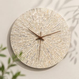 Peut inclure: Une horloge murale circulaire dotée d'un cadran en mosaïque composé de petites tuiles rectangulaires iridescentes disposées en rayons de soleil. L'horloge possède trois fines aiguilles en métal brun foncé centrées sur le cadran.
