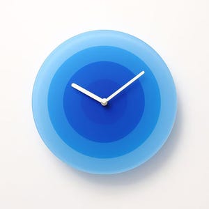 May include: A round wall clock with a gradient blue design. The clock face features concentric circles of varying shades of blue, with white clock hands. The clock is a modern design and is set against a white background.