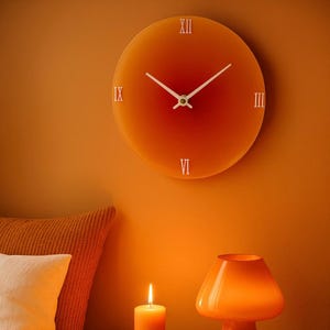 May include: A circular orange wall clock with white hands and Roman numerals. The clock is mounted on an orange wall. The clock face has a gradient effect, transitioning from a lighter orange to a deeper red in the centre.