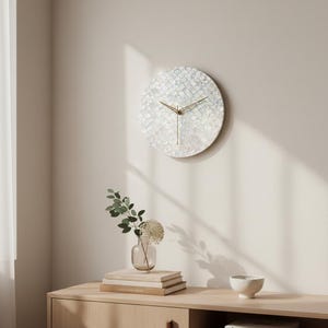 May include: A round wall clock with a mother-of-pearl face and gold-colored hands. The clock is mounted on a light-colored wall. The clock is a decorative item for home decor.