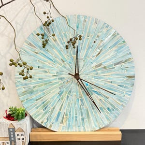 May include: A circular wall clock featuring a mosaic design of light blue and white glass tiles arranged in a sunburst pattern. The clock has three thin, dark brown metal hands at the centre and is supported by a rectangular wooden stand.