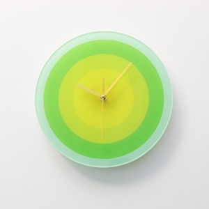 May include: A circular wall clock featuring a gradient of green and yellow concentric circles. The clock hands are gold-coloured. The clock face is made of glass. A decorative item for a home or office.