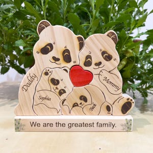 Peut inclure: Sculpture en bois représentant une famille de pandas avec un cœur rouge. Les mots "Daddy", "Mommy", "Sophia" et "Beck" sont gravés. La base indique "We are the greatest family."