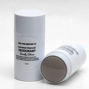 May include: Two white deodorant sticks with black text. One stick is upright, the other is on its side, revealing the charcoal deodorant. The label reads "Activated Charcoal Deodorant Barely Citrus" and lists ingredients.