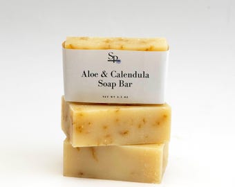 May include: A stack of three light yellow soap bars. The top bar has a white label that reads "Aloe & Calendula Soap Bar". The soap bars have a textured appearance and are stacked on top of each other. The net weight is 3.5 oz.