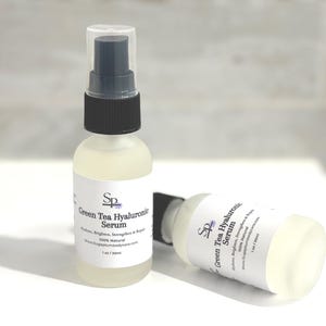 May include: Two clear glass spray bottles of Green Tea Hyaluronic Serum. The bottles have white labels with black text and a logo. The serum is a light green colour. The bottles are 30ml.
