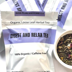 May include: A close-up of several silver pouches of "Digest and Relax Tea" from Suga Plum Bodycare LLC. The pouches have a purple band with the tea name and text that reads "100% Organic / Caffeine Free". A small bowl of loose leaf tea is visible.