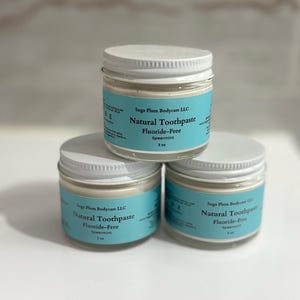 May include: Three clear glass jars with white screw-top lids, each containing natural toothpaste. The light blue labels read Suga Plum Bodycare LLC, Natural Toothpaste, Fluoride-Free, Spearmint, and 2 oz. The jars are arranged in a pyramid stack.