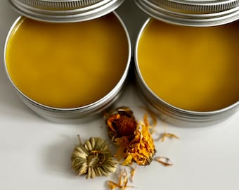May include: Two open, silver-colored tins filled with a bright yellow salve. Dried calendula flowers and buds are scattered near the tins. The labels on the tins read "Calendula Salve" and "Suga Plum Bodycare LLC."