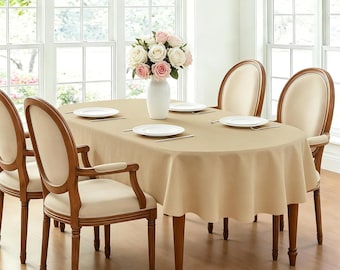 May include: An oval dining table covered with a solid beige tablecloth, set with four white plates and a central white vase containing a bouquet of pink and white roses, surrounded by four wooden chairs with beige upholstered seats and backs.