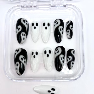 May include: A set of press-on nails in a clear plastic case. The nails feature a Halloween ghost design. Some nails are black with white ghost designs, while others are white with black ghost faces. One loose nail is also visible.