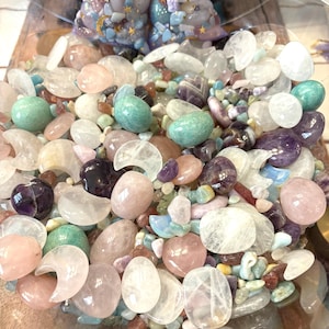 May include: A clear glass bowl filled with a variety of polished gemstones in shades of pink, green, purple, and clear. Two small, purple drawstring bags filled with similar stones are visible in the background. The stones vary in shape and size.