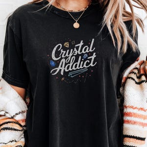 May include: A black short-sleeved t-shirt featuring the white text Crystal Addict in a script font. The design includes small illustrations of various crystals in blue, gold, and white, accented by tiny red star shapes scattered around the lettering.