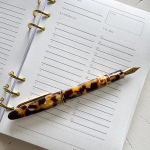 May include: A fountain pen with a brown and cream tortoiseshell pattern and a gold-colored nib resting on an open planner page. The planner features sections for day and date, top two priorities, a schedule, and a to-do list with checkboxes.