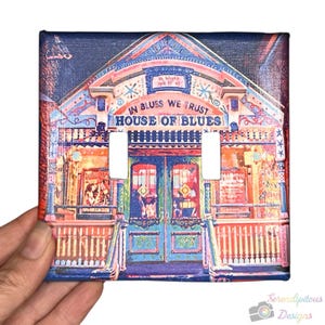Könnte beinhalten: Eine quadratische Lichtschalterabdeckung mit einer farbenfrohen Illustration des House of Blues. Das Design enthält den Text "In Blues We Trust" und "House of Blues" über den Schalteröffnungen. Das Kunstwerk ist in Blau-, Rot- und Orangetönen gehalten.