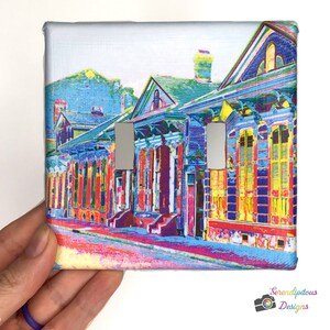 May include: A colorful light switch cover featuring a row of colorful houses with vibrant blue, yellow, red, and green accents. The houses have white trim and windows with shutters. The image is a digital illustration.