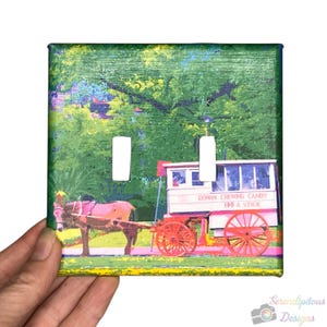 May include: A square light switch cover with a vibrant image of a horse-drawn candy cart. The cart is red and white, with the text "ROMAN CHEWING CANDY 100 A STICK". The background is a green, impressionistic landscape.