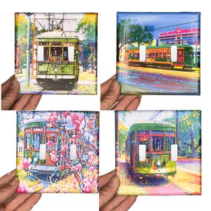 May include: Four decorative light switch covers with vibrant illustrations of vintage trams. Each cover presents a unique artistic depiction of the trams, with diverse backgrounds and floral details. The covers are held by hands.