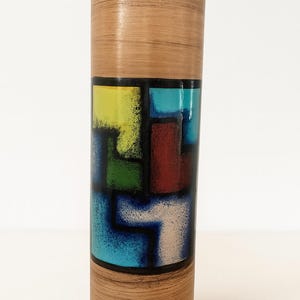 Op de afbeelding: Een hoge, cilindervormige vaas met een getextureerde, lichtbruine buitenkant. Een band van kleurrijke, geometrische vormen in geel, groen, rood, blauw en wit is gecentreerd op de vaas. De vaas heeft een donkerbruine rand.