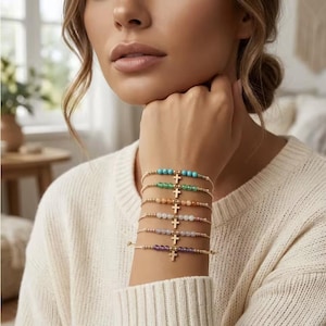 Puede incluir: Una pila de seis pulseras delicadas en la muñeca. Cada pulsera cuenta con un cordón fino, pequeñas cuentas doradas y un dije de cruz central en tono dorado. Las cuentas de cada pulsera son de un color diferente: turquesa, verde, naranja, blanco, morado claro y morado oscuro.
