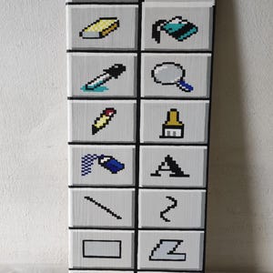 Könnte beinhalten: Vertikale Holztafel mit 16 pixeligen Symbolen. Die Symbole sind in Weiß, Schwarz, Gelb und Blau gehalten und in einem Raster angeordnet. Die Symbole stellen verschiedene Formen und Werkzeuge dar, die an frühe Computergrafiken erinnern.