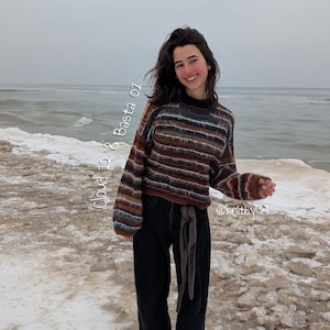 Könnte beinhalten: Eine Person trägt einen gestreiften braunen, blauen und burgunderfarbenen Pullover mit schwarzer Hose und schwarzen Schuhen. Der Pullover hat einen hohen Kragen und lange Ärmel. Der Text "Cloud 12 & Basta 01" befindet sich auf der linken Seite des Bildes.