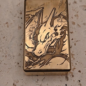 May include: A gold-colored metal lighter with an engraved design of a dragon-like creature. The lighter has a rectangular shape and a hinged lid. The design is detailed and features flames and sharp edges. The text "ULTIMATE JUSTICE" is visible on the bottom.