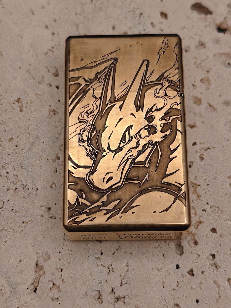 May include: A gold-colored metal lighter with an engraved design of a dragon-like creature. The lighter has a rectangular shape and a hinged lid. The design is detailed and features flames and sharp edges. The text "ULTIMATE JUSTICE" is visible on the bottom.