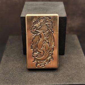 May include: A brass-colored lighter with a detailed dragon design. The dragon is depicted with wings and is surrounded by swirling patterns. The lighter is rectangular and has a hinged lid. The design is embossed on the front of the lighter.