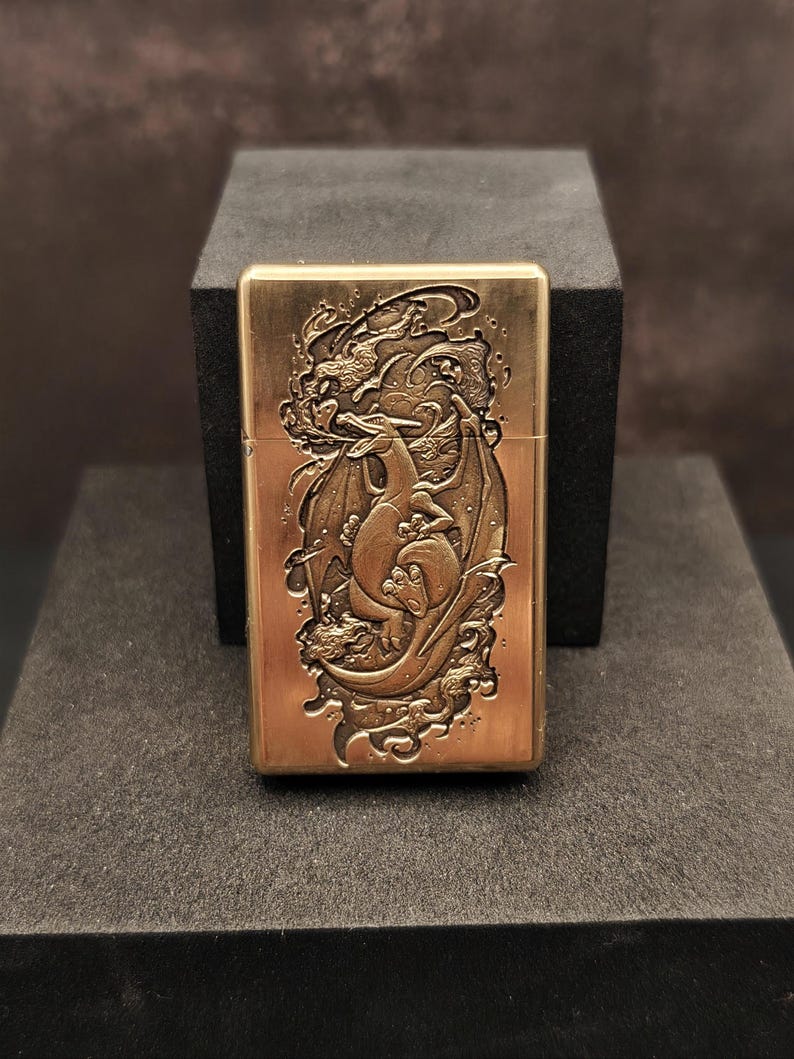 May include: A brass-colored lighter with a detailed dragon design. The dragon is depicted with wings and is surrounded by swirling patterns. The lighter is rectangular and has a hinged lid. The design is embossed on the front of the lighter.