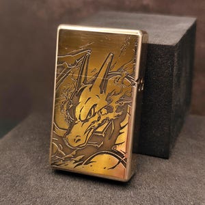 May include: A brass-colored lighter with a detailed engraved design of a dragon. The dragon is depicted with flames and intricate detailing. The lighter is rectangular and has a hinged lid.