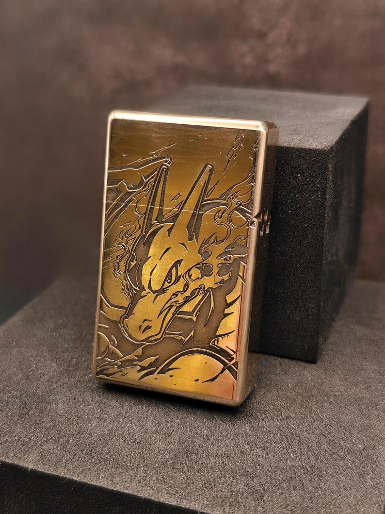 May include: A brass-colored lighter with a detailed engraved design of a dragon. The dragon is depicted with flames and intricate detailing. The lighter is rectangular and has a hinged lid.
