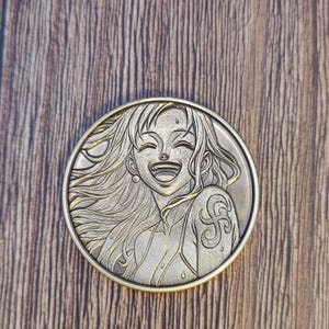Può includere: Una moneta rotonda color bronzo raffigurante un personaggio anime sorridente con lunghi capelli fluenti. La moneta ha un design a rilievo dettagliato e un bordo decorativo. Lo sfondo è una superficie di legno.