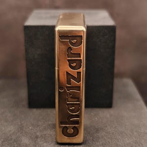 May include: A brass-colored lighter with the word "Charizard" engraved vertically. The lighter has a rectangular shape with rounded edges and a shiny finish. The background is a dark gray cube.