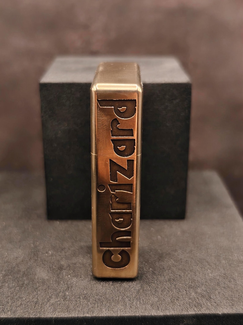 May include: A brass-colored lighter with the word "Charizard" engraved vertically. The lighter has a rectangular shape with rounded edges and a shiny finish. The background is a dark gray cube.