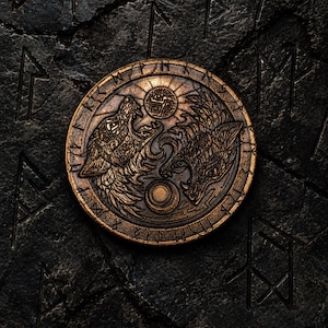 Puede incluir: Un medallón circular de tono bronce que presenta dos cabezas de lobo detalladas frente a frente en una composición estilo yin-yang. El diseño incluye un motivo central de sol y luna, rodeado por un borde exterior grabado con caracteres rúnicos.