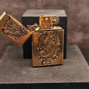 May include: A brass-colored lighter with an open lid, revealing an engraved dragon design. The lighter is rectangular with rounded edges and a hinged lid. The dragon is depicted in a detailed, artistic style, with intricate scales and wings.