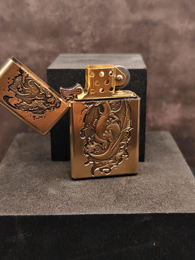 May include: A brass-colored lighter with an open lid, revealing an engraved dragon design. The lighter is rectangular with rounded edges and a hinged lid. The dragon is depicted in a detailed, artistic style, with intricate scales and wings.