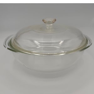 May include: A clear glass casserole dish with a matching lid. The round dish features two integrated side handles and a circular knob on the center of the lid. The glass is transparent, smooth, and has a polished finish throughout the entire piece.