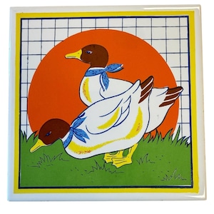 Puede incluir: Un azulejo de cerámica cuadrado con una ilustración gráfica de dos patos blancos con cabezas marrones y pañuelos azules. Los patos están sobre hierba verde frente a un gran círculo naranja y un fondo de cuadrícula blanca con borde amarillo.